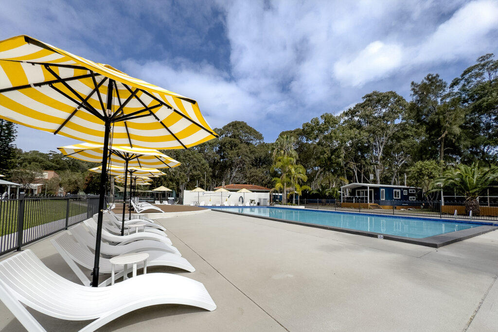 An outdoor swimming pool surrounded by lounge chairs and large, yellow-and-white striped umbrellas evokes the relaxing vibes of Kioloa Beach. Bordered by trees and greenery, the sky above is partly cloudy, providing a serene and inviting atmosphere.