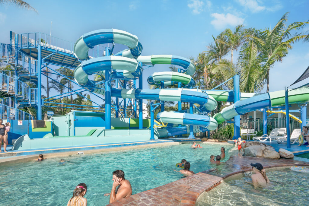 A water park scene features several people enjoying a pool. In the background, there are blue and green waterslides. Palm trees and a clear sky complete the lively, summery atmosphere.