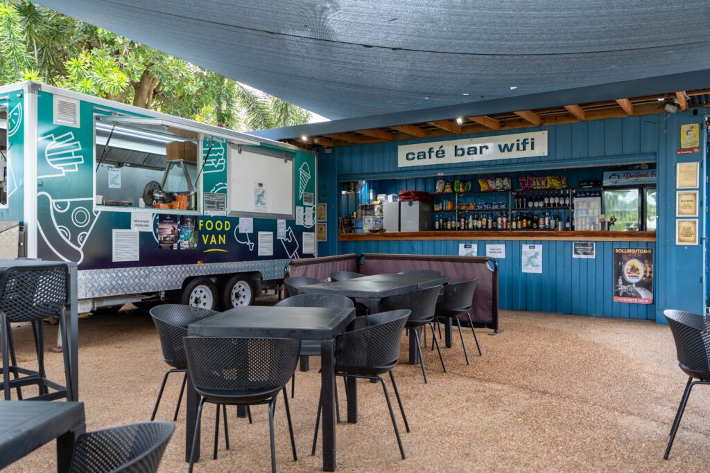 Outdoor café area with black tables and chairs situated in front of a Rollingstone food truck. The truck features a green and white design with "Food Van" written on it. A wooden bar with shelves of drinks and a "café bar wifi" sign is under a blue awning.