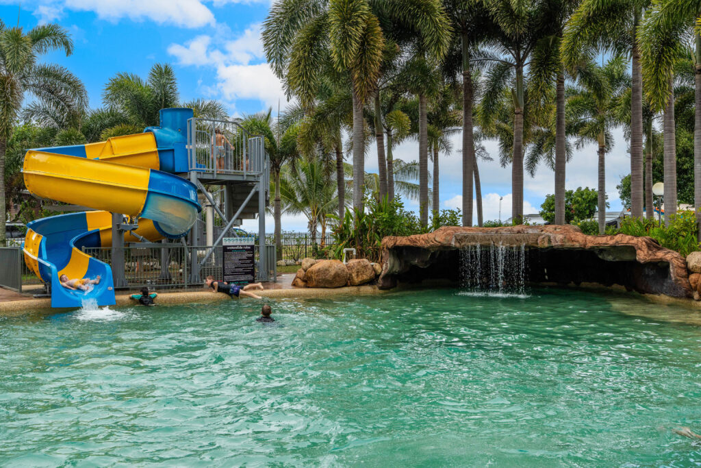 A tropical pool area with palm trees, a yellow and blue water slide, and a rock formation with a waterfall. Several people are enjoying the pool, and a person is sliding down the water slide into the pool. The sky is partly cloudy.