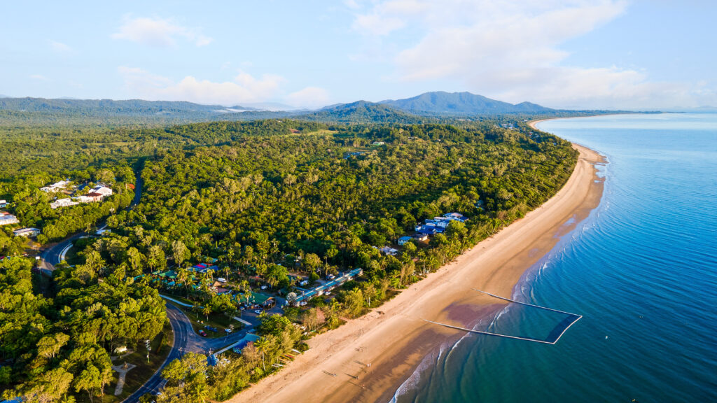 Aerial view of a scenic coastal area, featuring the long sandy stretch of Mission Beach lined by lush green forests. A few buildings and houses are scattered along the shoreline. The coastline curves gently, bordered by calm blue waters on one side and dense greenery on the other.