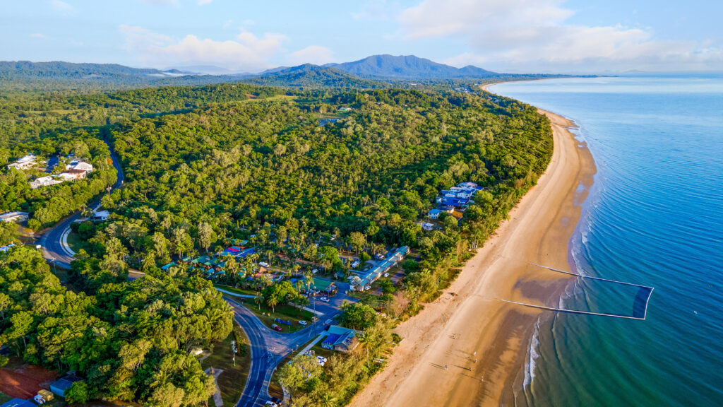 Aerial view of Mission Beach, a coastal area with a long sandy beach bordered by lush green forest. Beachfront holiday park and roads are visible near the shore. The calm blue sea stretches out to the right, and a mountainscape is visible in the background.