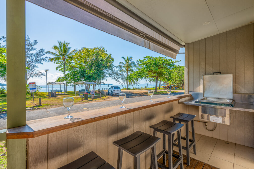 An outdoor kitchen area with bar stools and a counter overlooking a scenic view of Mission Beach's park, dotted with palm trees, picnic tables, and a body of water. Two wine glasses are placed on the counter, and a grill is built into the setup.