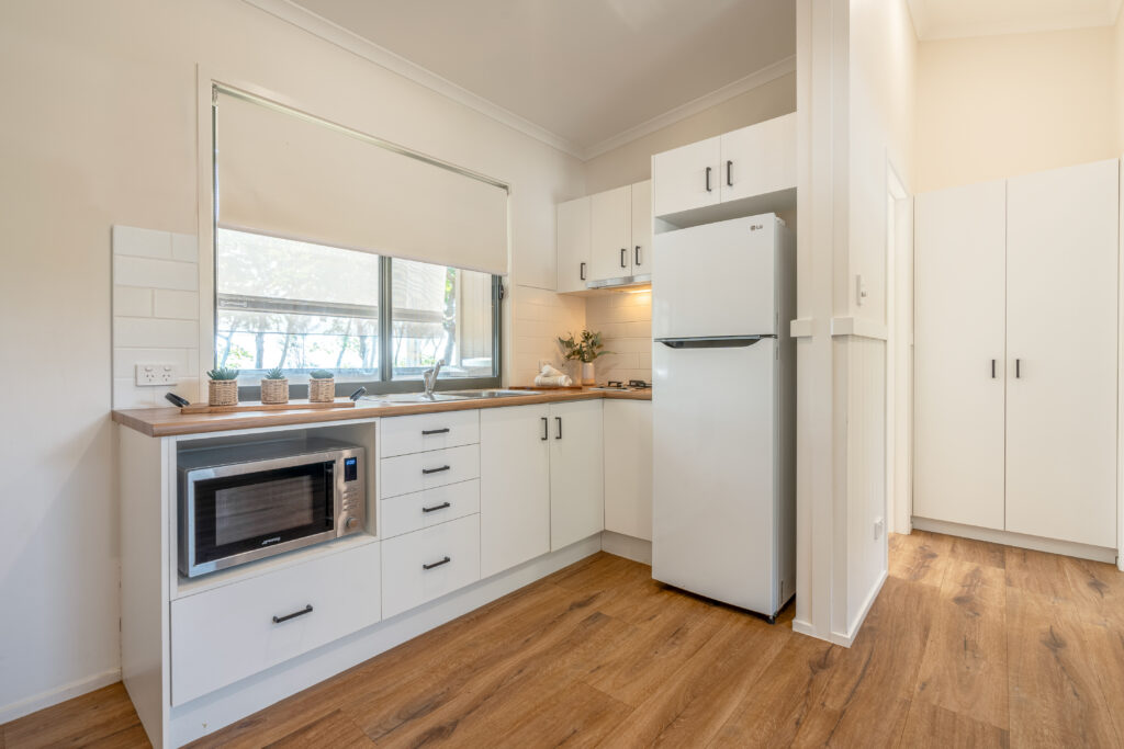 A modern, compact kitchen in Mission Beach features white cabinetry, a wooden countertop, and stainless steel appliances. The kitchen includes a microwave, sink, electric stovetop, and tall refrigerator. Light streams in through a window with a roller blind, reflecting off the wooden floor.