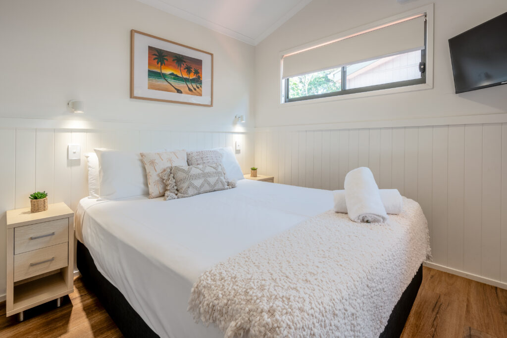 A cosy bedroom with a neatly made double bed, adorned with white and patterned pillows. Folded towels rest on a beige throw at the foot of the bed. The room features a bedside table, a scenic painting of Mission Beach, a wall-mounted TV, and a small window.