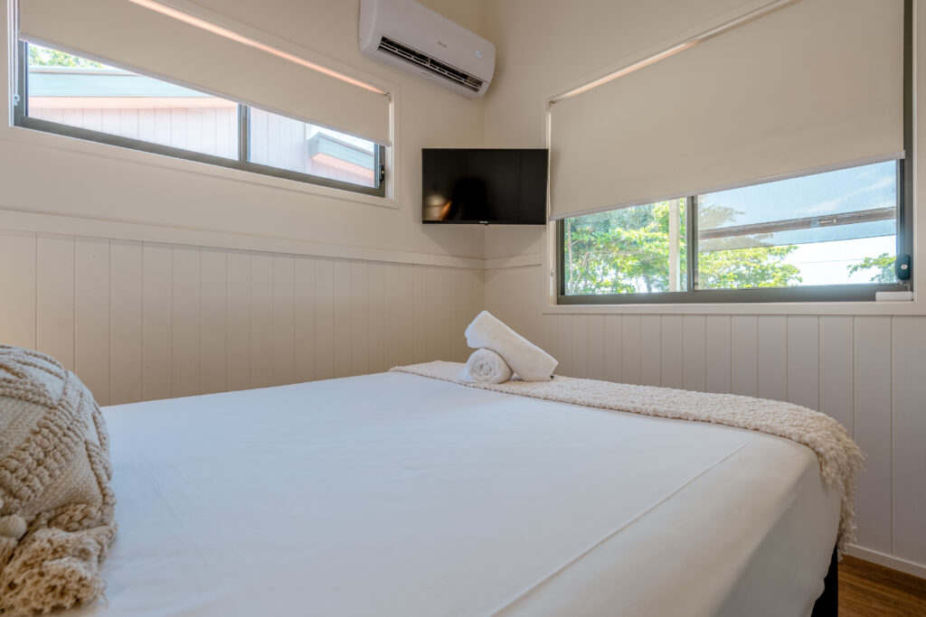 A bright bedroom with white walls in Mission Beach features a neatly made bed with a white knitted throw and a towel folded on top. Two windows with shades allow natural light in. An air conditioner is mounted on the wall above a small, flat-screen TV beside the window.