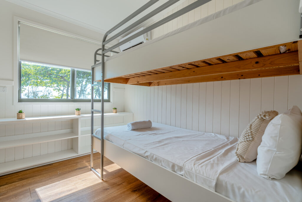 A bright room with white walls features a bunk bed. The lower bed is neatly made with white linens and a rolled towel. A large window, offering views of Mission Beach and lush greenery, allows natural light to fill the space. Built-in shelves beneath the window hold small potted plants.