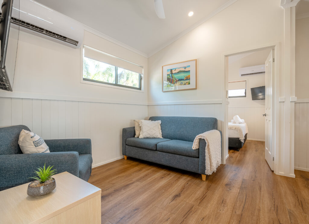 A cosy living area with a blue sofa, matching armchair, and a light wood coffee table with a small potted plant. The room features wooden flooring and white walls, evoking the serene vibes of Mission Beach. A doorway leads to a bedroom with a double bed. A wall-mounted TV and air conditioner are visible.