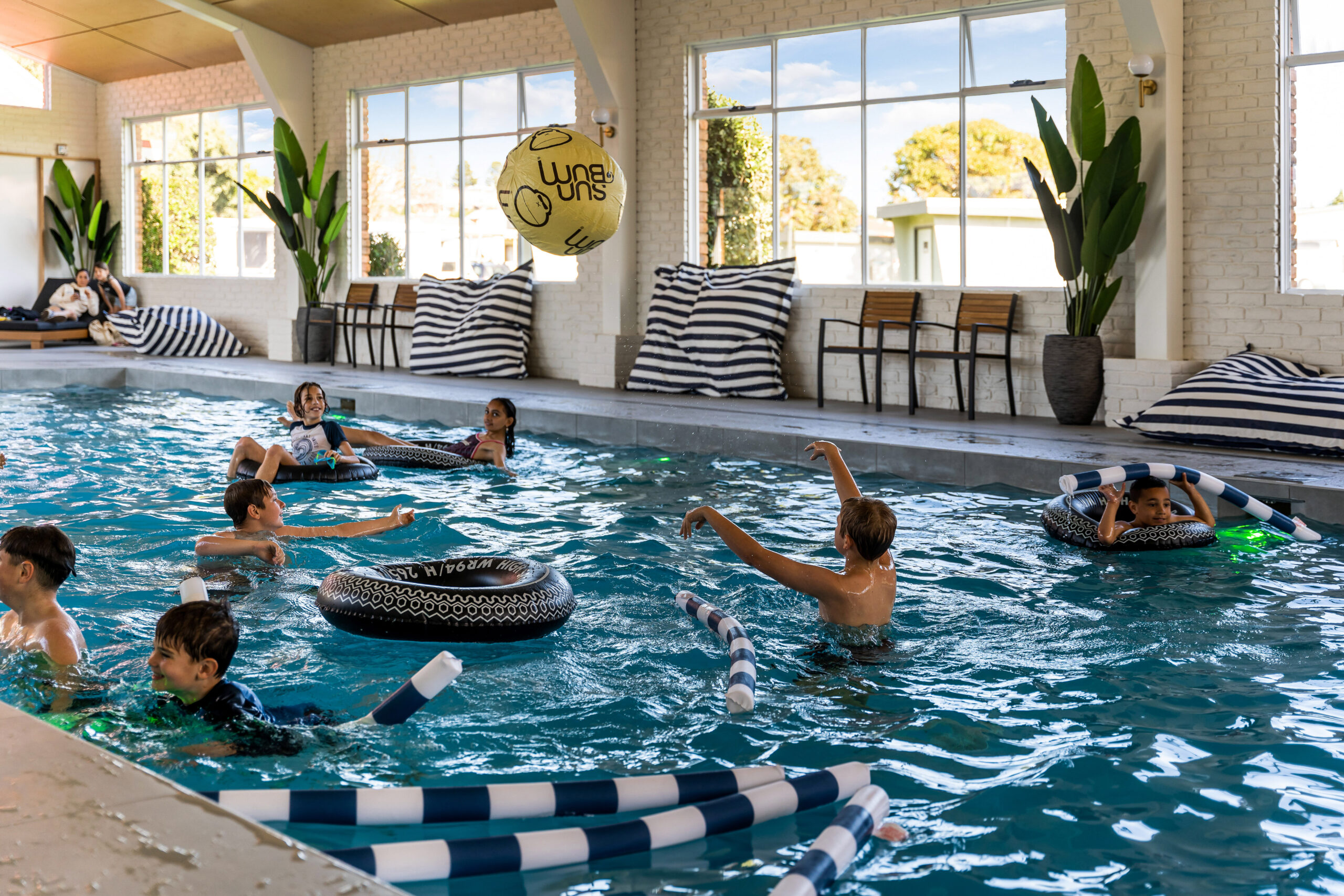 A group of children is playing in an indoor swimming pool. Some are floating on inflatable rings while others are in the water. They are tossing a large inflatable ball. The pool area is lined with striped cushions, plants, and large windows showcasing an outdoor scene.