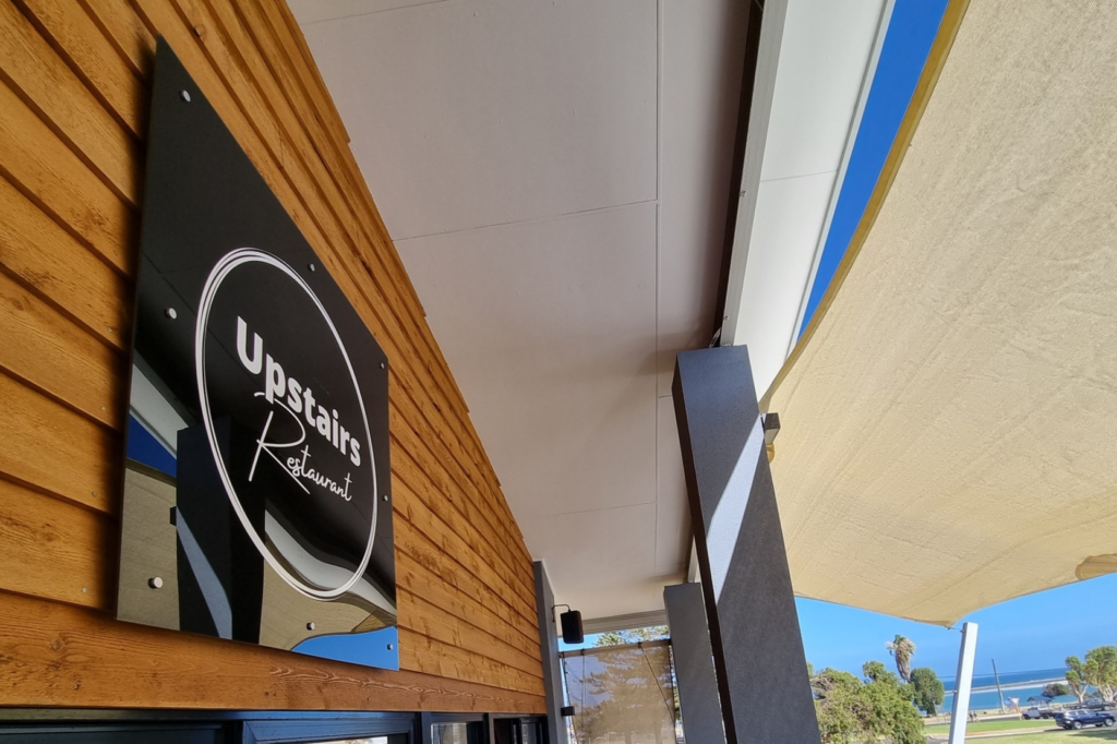 Outdoor seating area of a restaurant with tables and chairs, partially shaded by a sail canopy. A sign reads "Upstairs Restaurant." The restaurant overlooks a scenic view of greenery and the ocean in the background.