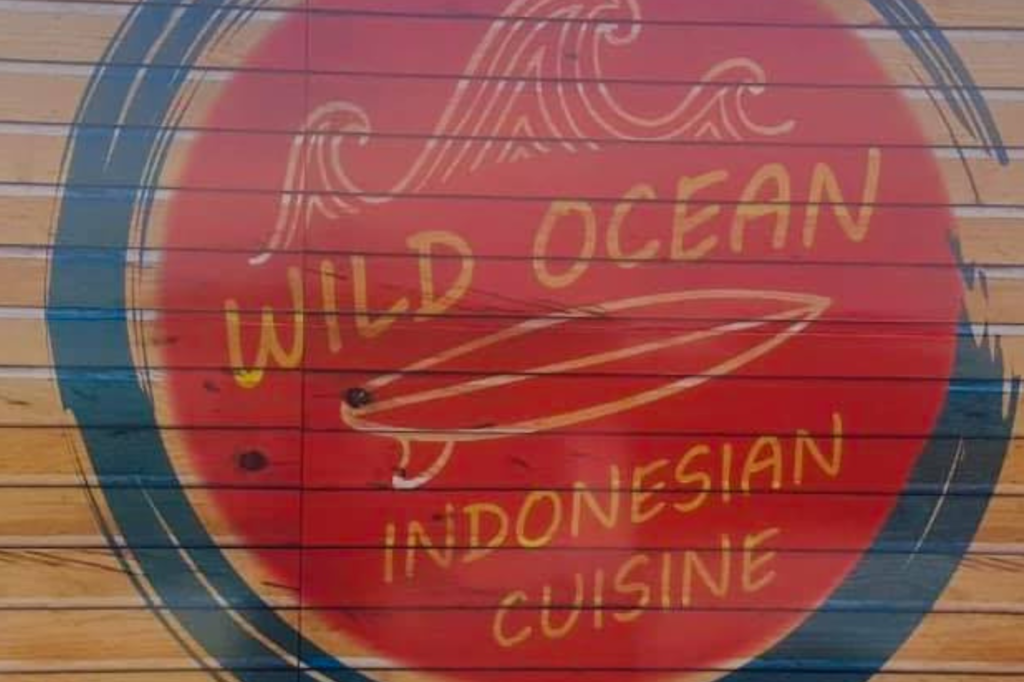 A wooden wall features a large red circle with a wave design and a surfboard illustration. The text within the circle reads "Wild Ocean Indonesian Cuisine" in yellow. A dark blue border surrounds the circle.