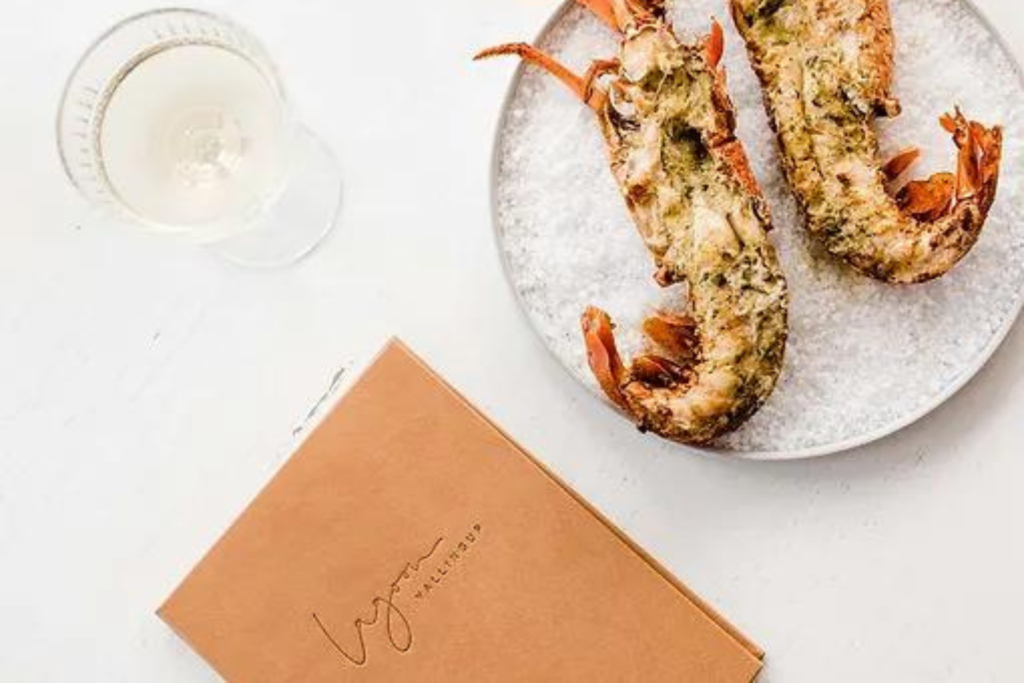 A plate featuring two seasoned and grilled lobsters is placed on a white surface. A glass of white wine sits beside the plate, and a brown menu titled "Lynn at The Whaler's Inn" is partially visible at the bottom of the image.