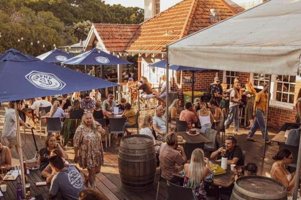 People sit and converse at an outdoor restaurant with wooden barrels as tables. The area is shaded by large blue umbrellas, and a light-colored brick building with a tiled roof is in the background. String lights and trees add to the warm, bustling atmosphere.