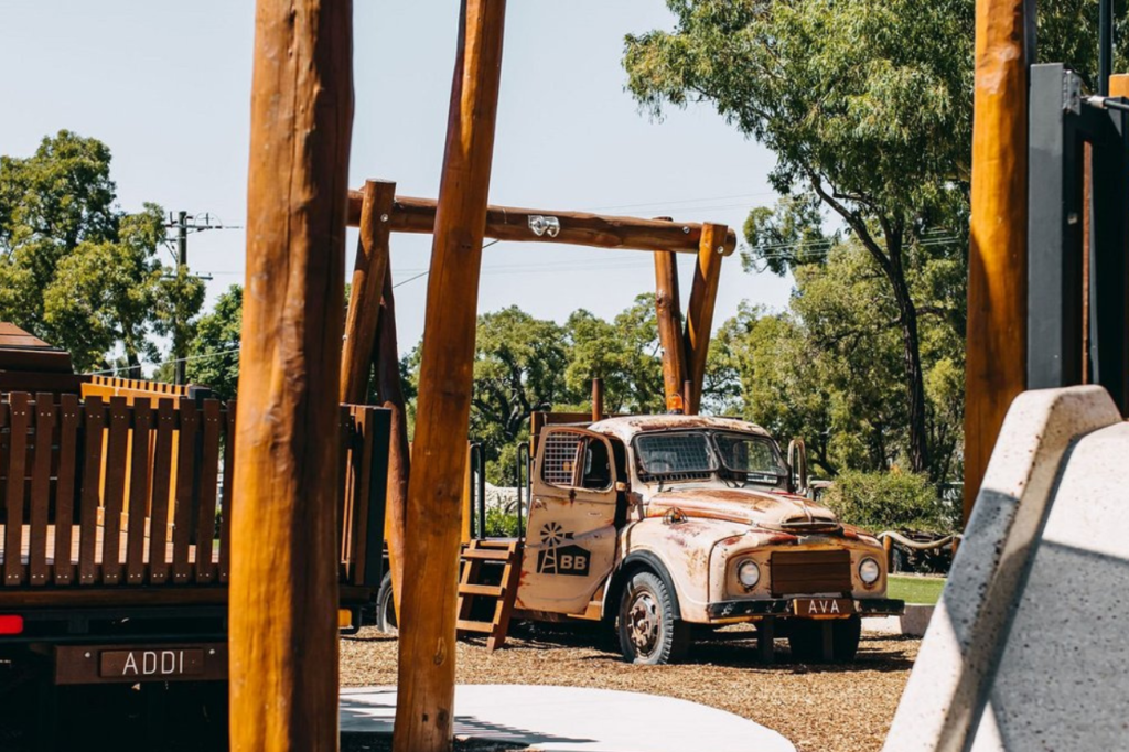 A playground with wooden structures including a swing set and an old brown truck repurposed as a playhouse. The area has wood chip flooring and is surrounded by trees and greenery. A concrete path is visible, and a slide is partially seen on the right.