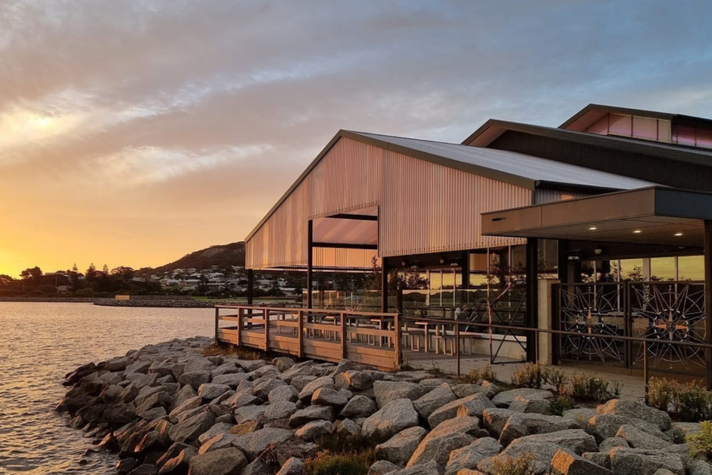 A modern, glass-fronted building with a gabled roof sits on a rocky shoreline, overlooking a calm body of water at sunset. The sky is tinged with hues of orange and pink, casting a warm glow on the structure. A hill with houses is visible in the background.