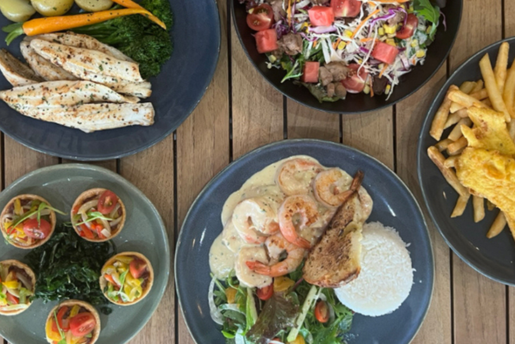 A top view of a wooden table with five plates of food: grilled chicken with potatoes and carrots, a mixed salad with watermelon chunks, a plate of fries with fried fish, a dish with rice, grilled shrimp, and grilled fish, and a plate of small, colorful appetizers.