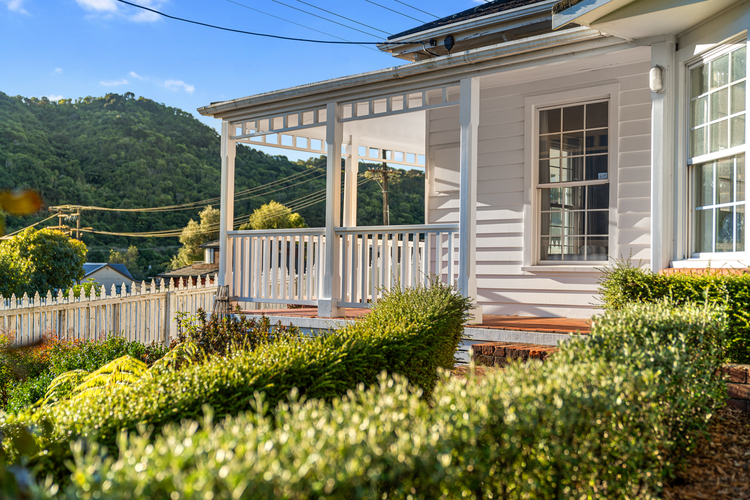 A white house with a wraparound porch and white railing sits near a garden with neatly trimmed hedges. The background features green, tree-covered hills under a clear blue sky. A white picket fence lines the property, creating the perfect scene for discovering things to do in Papamoa.