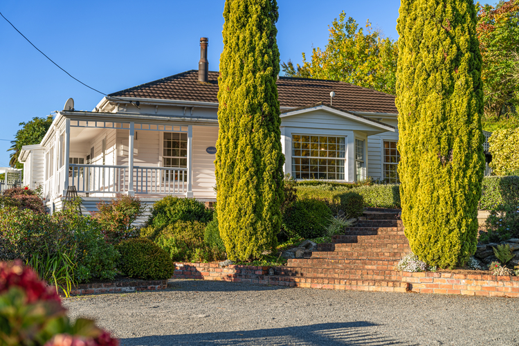 A charming white house with a wraparound porch is surrounded by lush greenery. Two tall, narrow evergreen trees flank the brick stairway leading to the front entrance. Reminiscent of the tranquil setting found at Tasman Holiday Parks - Picton, the clear blue sky emphasizes the home's serene and welcoming atmosphere.