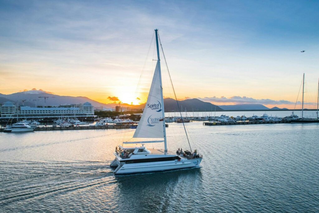 A catamaran sails across calm waters under a clear sky during sunset. The vessel's sail is unfurled, and people are visible onboard. In the background, the marina in Cairns is dotted with docked boats, and mountains are silhouetted against the setting sun.