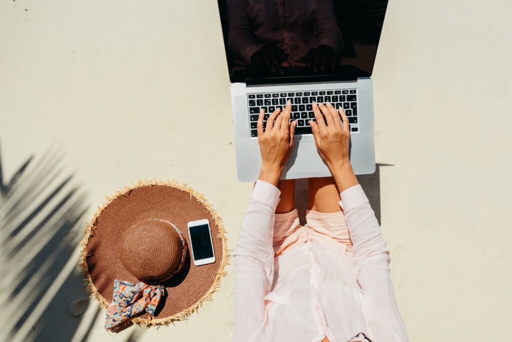 A digital nomad is sitting on the sand with a laptop on their lap, typing. Nearby are a straw hat with a patterned ribbon and a smartphone placed on the sand. Shadows of palm leaves are visible on the left side.