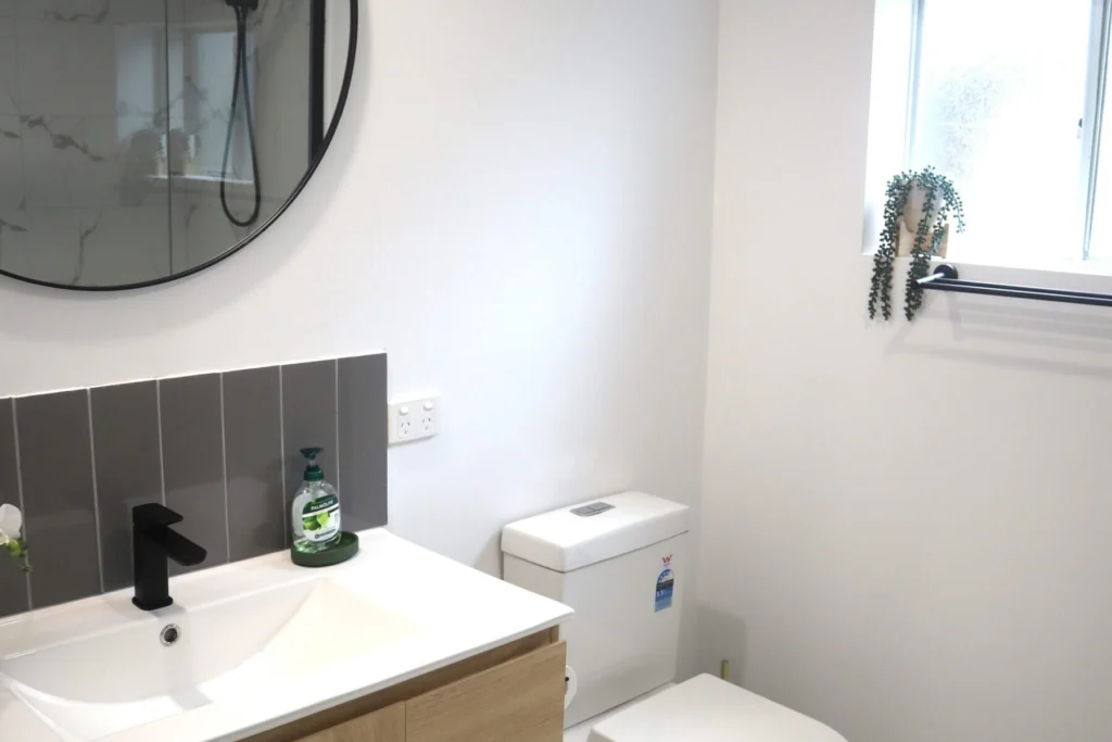 A modern bathroom features a wooden vanity with a white sink, black faucet, and round mirror above. A toilet is next to the vanity. The wall behind the sink has vertical grey tiles. A small window with a potted plant on the sill brings natural light into the space.