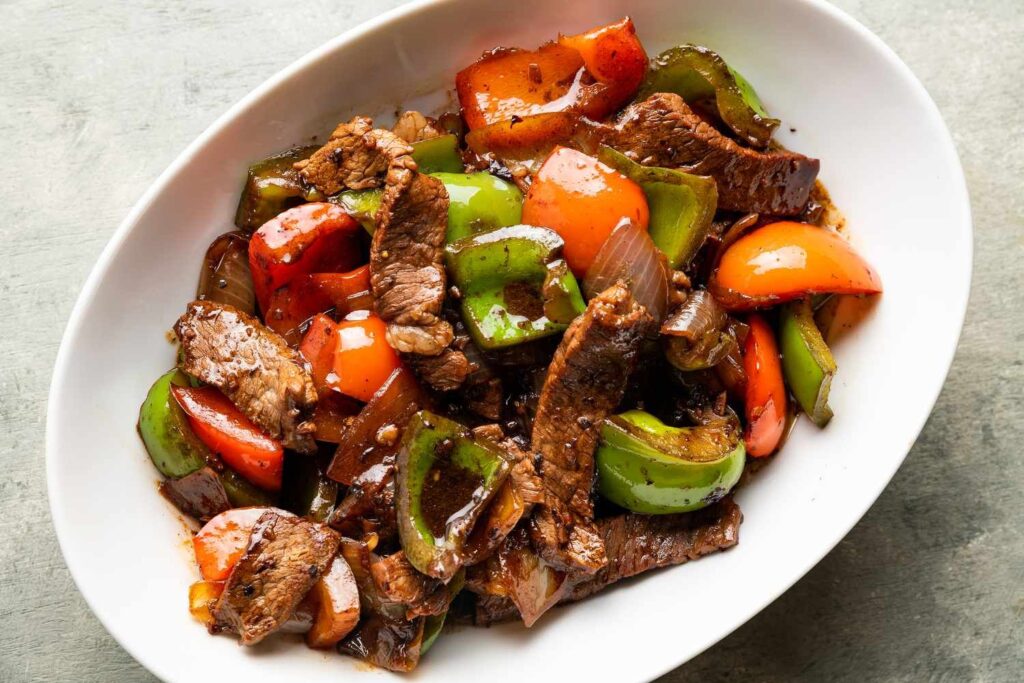 A white dish filled with a stir-fry of sliced beef, green and red bell peppers, and onions, all coated in a glossy sauce. The collective ingredients create a colorful and appetizing presentation.