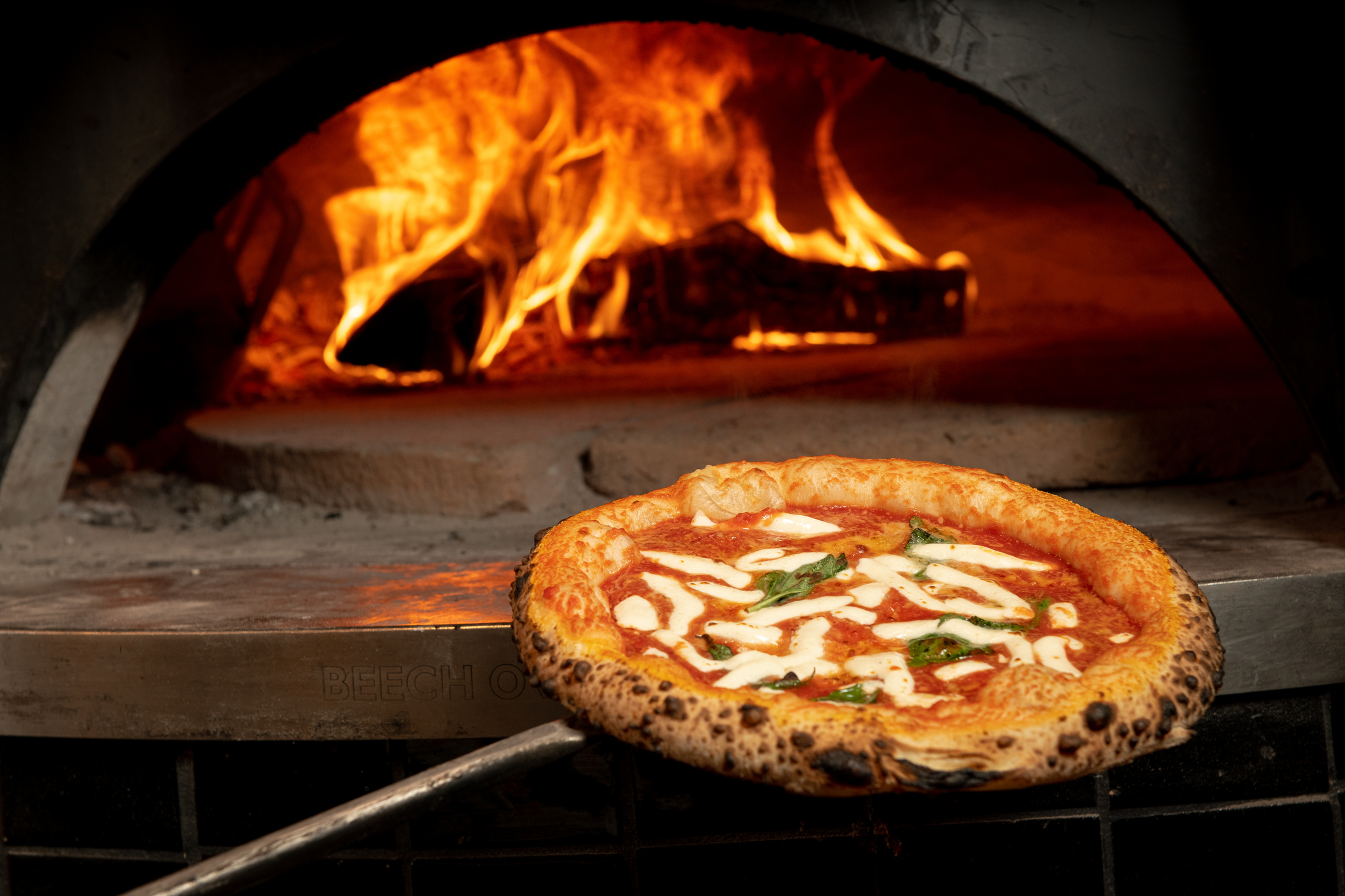 A wood-fired oven with a roaring flame in the background. In the foreground, a margherita pizza on a wooden peel, topped with tomato sauce, melted mozzarella cheese, and fresh basil, is being pulled out of the oven.