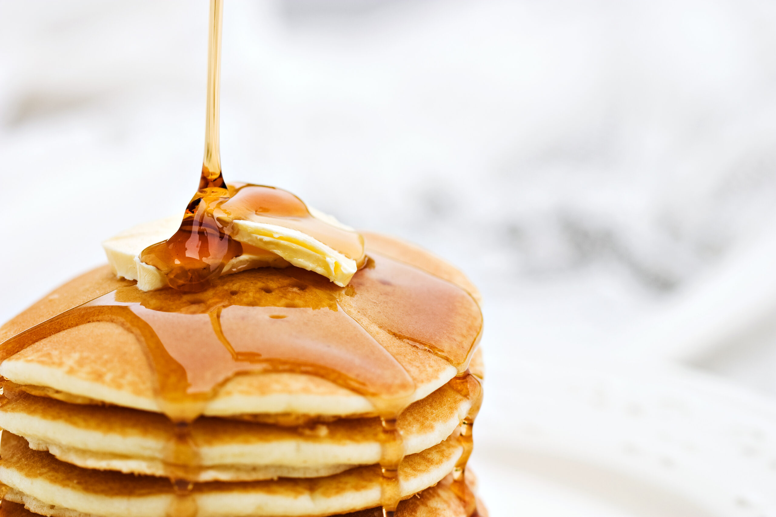 A close-up image of a stack of golden brown pancakes being drizzled with maple syrup. The pancakes have a pat of melting butter on top, and the syrup is captured mid-pour, glistening in the light. The background is softly blurred.