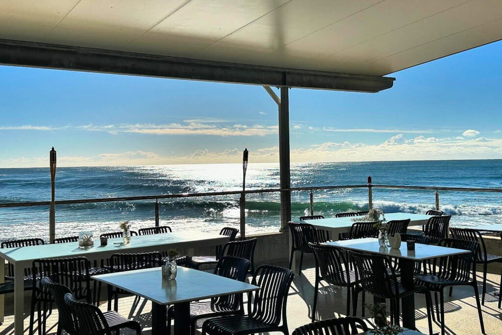 An outdoor seaside restaurant with several empty tables and chairs under a covered patio. The sun shines brightly, reflecting off the ocean waves in the background. Small flower arrangements adorn the tables, and tall torches line the edge of the railing.