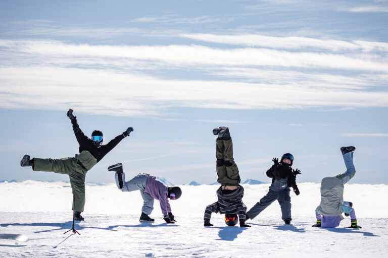 Five people are performing acrobatic poses on a snowy landscape with a clear sky and mountains in the background. Embracing the spirit of a winter adventure, they are dressed in jackets, pants, and gloves. One person is doing a handstand, while others strike varied stances.