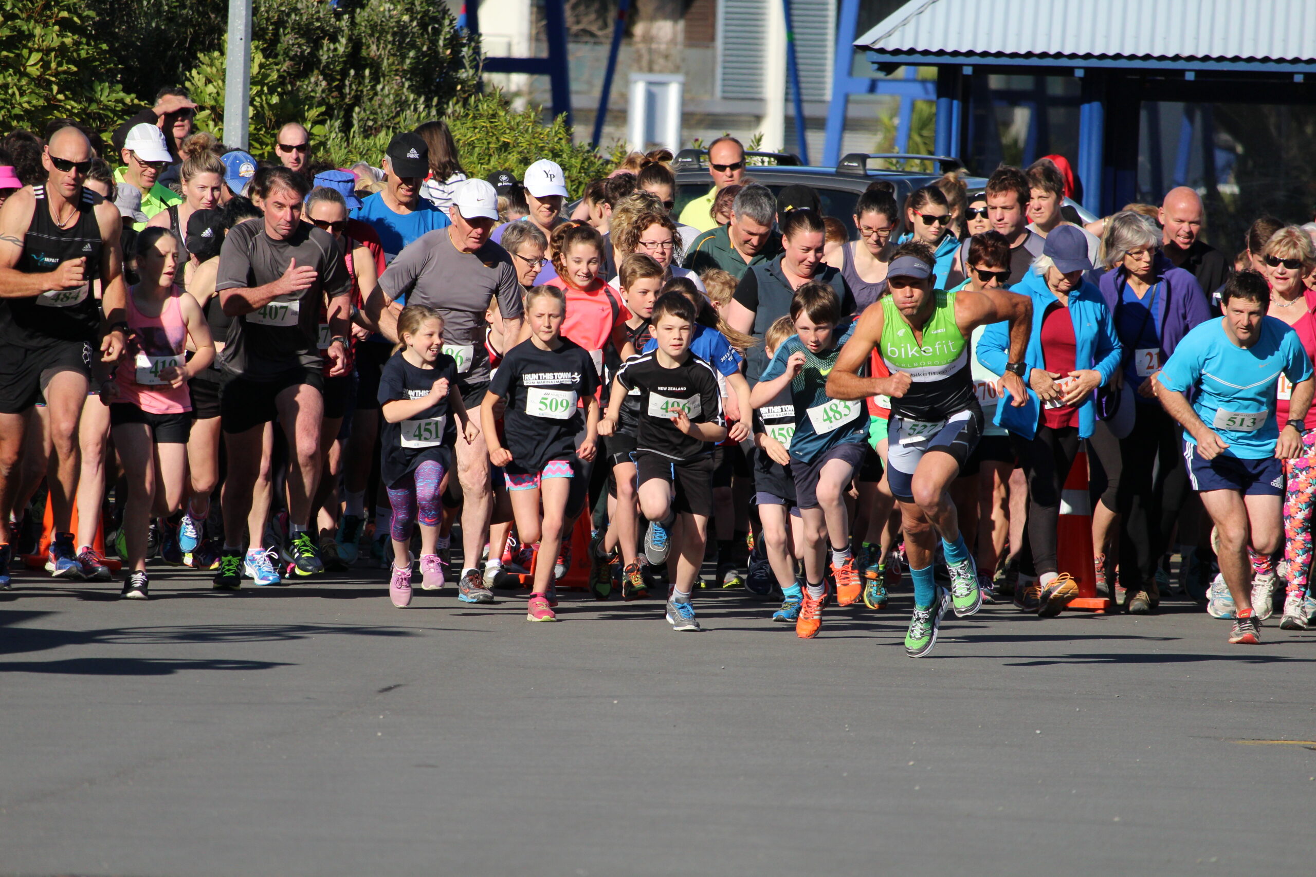 A group of runners of various ages, including men, women, and children, are starting a race near some attractions near Picton in spring. They are all wearing athletic gear and race bibs with numbers. The background includes spectators, trees, and buildings. Everyone appears focused and energetic.