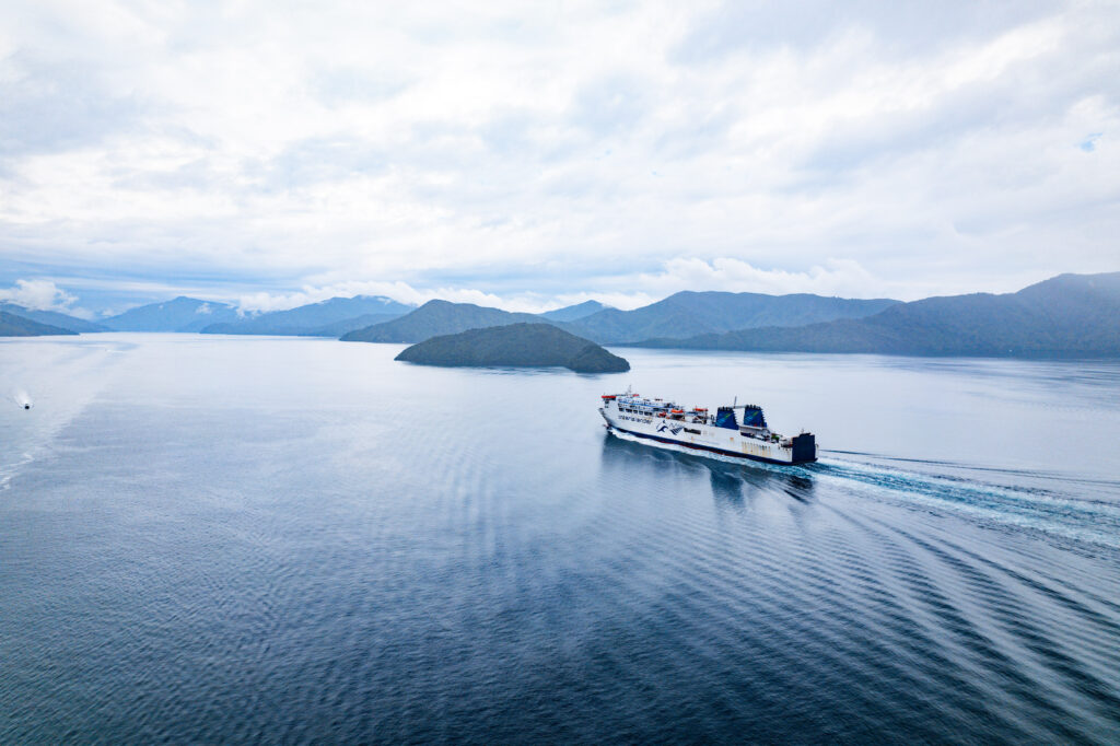 A large ferry sails across calm, blue waters amidst mountainous, green islands under a cloudy sky. The wake of the ferry creates ripples in the water, highlighting the serene and expansive landscape, perfect for exploring attractions near Picton.