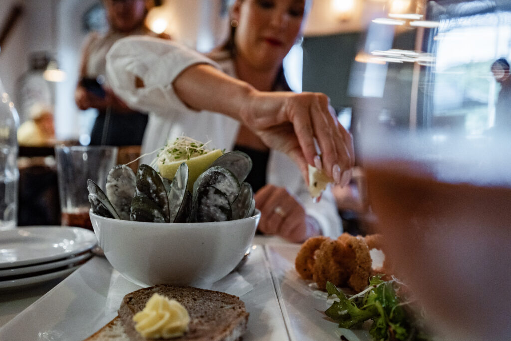 A person reaches to pick up a piece of food from a plate on a table. In the foreground, there is a bowl of mussels, a slice of bread with butter, and some breaded morsels, evoking the culinary delights one might find exploring attractions near Picton. Other dining elements are partially visible in the background.