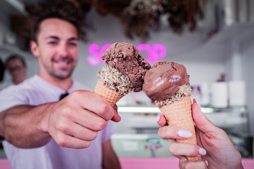 Two people holding ice cream cones, each topped with two scoops of chocolate ice cream. The person on the left is smiling and wearing a light-colored shirt. The background includes a neon sign reading "SCOOP" and ice cream shop decor, reminiscent of the quaint attractions near Picton.