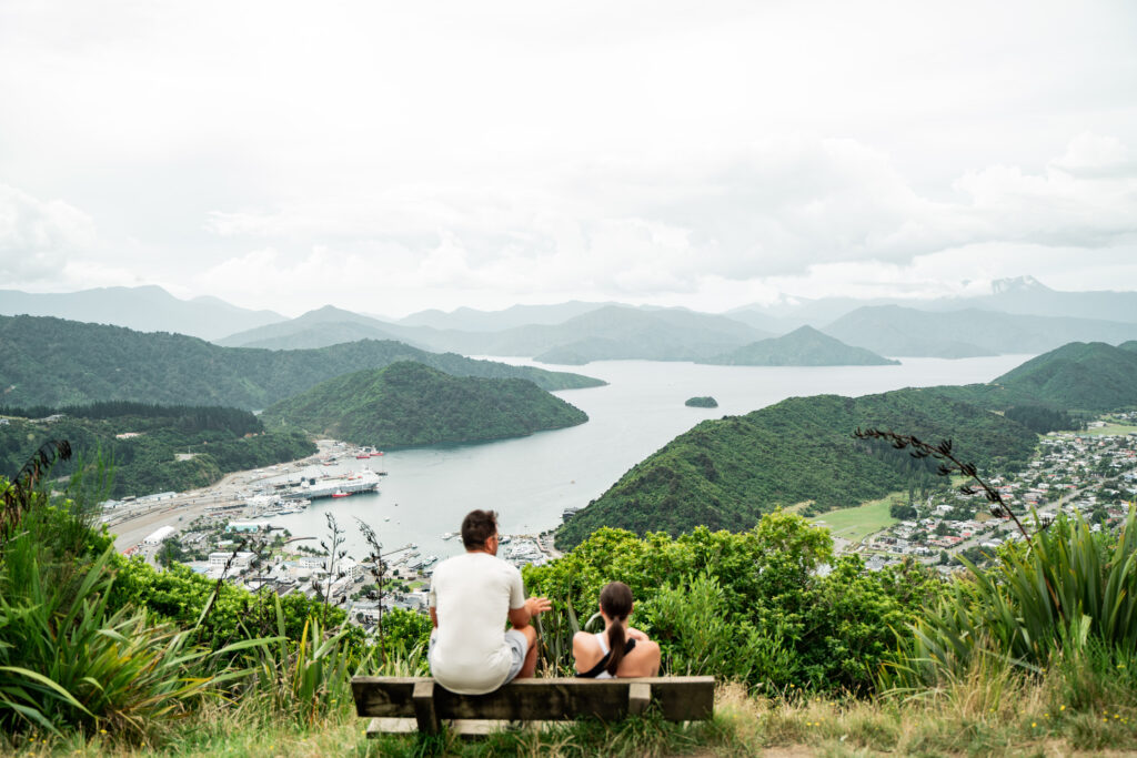 Two people sit on a wooden bench overlooking a scenic view of Picton in spring, with green hills and mountains surrounding the bay. The sky is cloudy, and boats are docked at a marina. Verdant foliage frames the foreground, with a small town visible to the right.