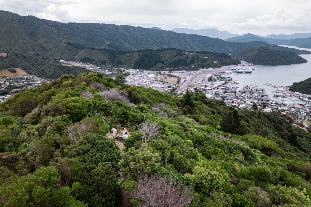 Two people sit on a small clearing atop a lush, green hill. Below them, a coastal town with boats docked in a harbor stretches along the shoreline. Surrounding mountains and cloudy skies form the backdrop in this scenic view of attractions near Picton.