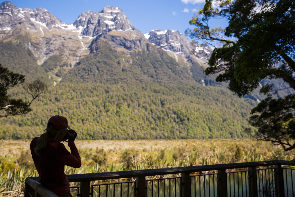 A person stands on a wooden deck, observing natural water wonders, using binoculars to observe the scenic view of a mountain range covered in snow. The foreground features lush greenery and a forest, with a clear blue sky overhead. The scene is peaceful and nature-oriented.