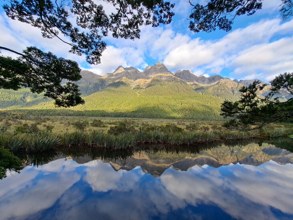 A serene landscape featuring a clear river reflecting nearby mountains and a partly cloudy sky. Lush greenery lines the riverbanks and the mountains, with trees framing the scene from above.