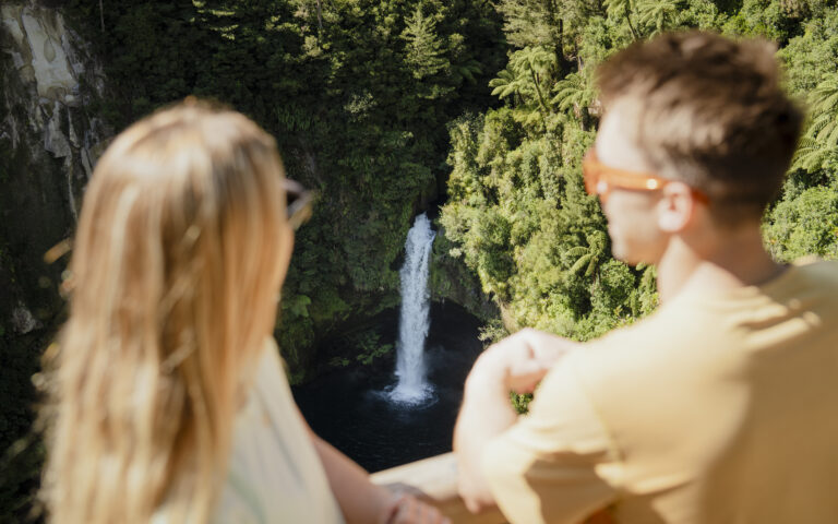 A man and woman with blurred faces in the foreground look at a waterfall cascading into a lush, green forest.