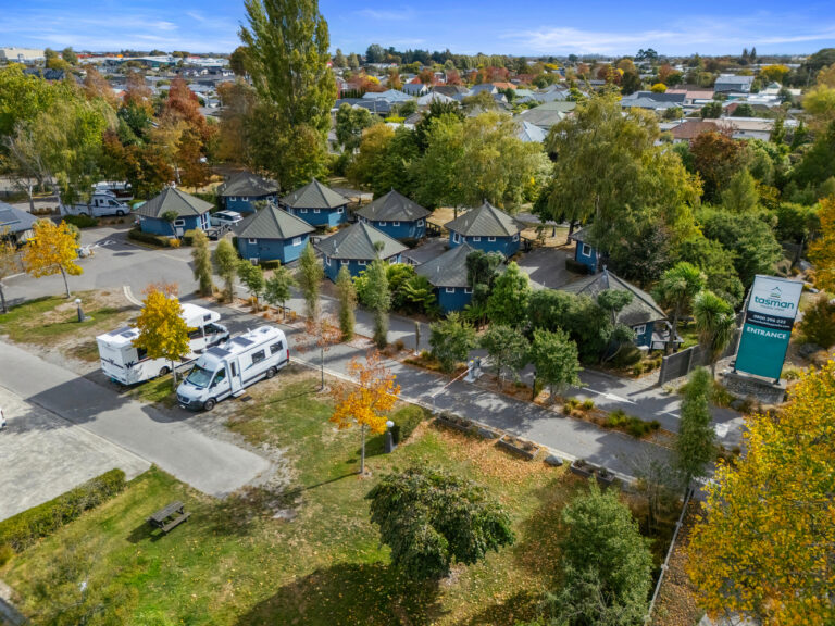 Aerial view of a caravan park featuring several teal-colored, hexagonal cabins surrounded by trees with autumn foliage. Two white campervans are parked in an open area near the cabins. A sign at the entrance reads "Tasman Holiday Parks." Houses are visible in the background.