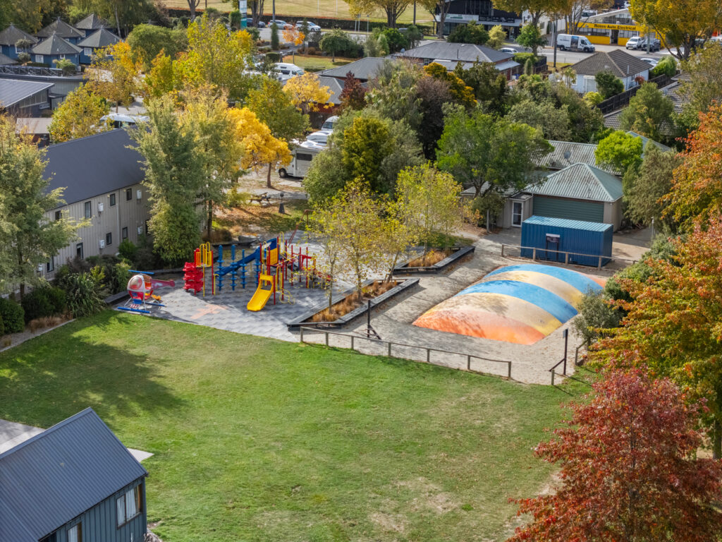 Aerial view of a playground in a park. The playground features colorful climbing structures, slides, swings, and a large inflatable air cushion. Surrounding the playground are trees with autumn foliage, a green lawn, nearby buildings, and parked cars.