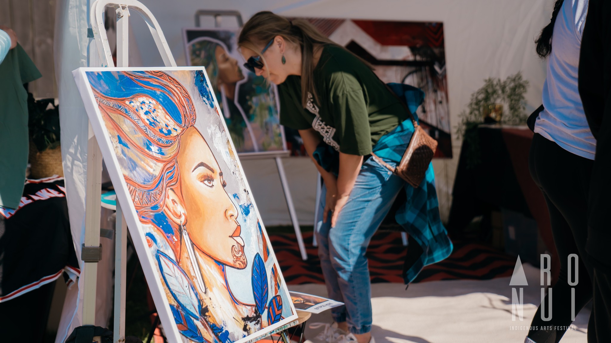 A woman wearing sunglasses and a green shirt with white text leans towards a colorful, detailed painting of a woman's profile displayed on an easel. The art exhibition has various other artworks in the background and is bright with sunlight. The logo "ARONUI" is visible.