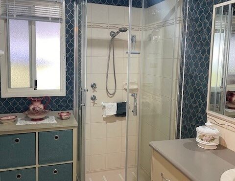 A bathroom with a glass shower enclosure, blue patterned wallpaper, and beige tiles. The shower area has silver fixtures, a showerhead, and a towel hanging inside. A sink with a counter, mirror, and drawers is to the right. A window with blinds is on the left.