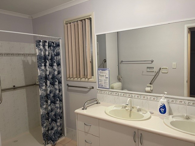 A bathroom with a double sink and large mirror above. A hand soap dispenser is on the counter. To the left is a walk-in shower with a blue and white floral curtain. Safety bars are installed near the shower and sinks. Vertical blinds cover the window.