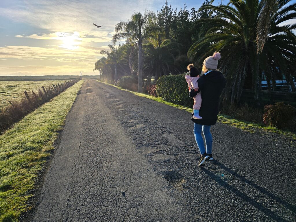 A person in a black coat and pink beanie is carrying a small child dressed in a pink outfit. They are walking down a rural road lined with palm trees, enjoying family-friendly spring activities in Miranda under a bright sky with the sun setting or rising. A bird is flying in the sky.