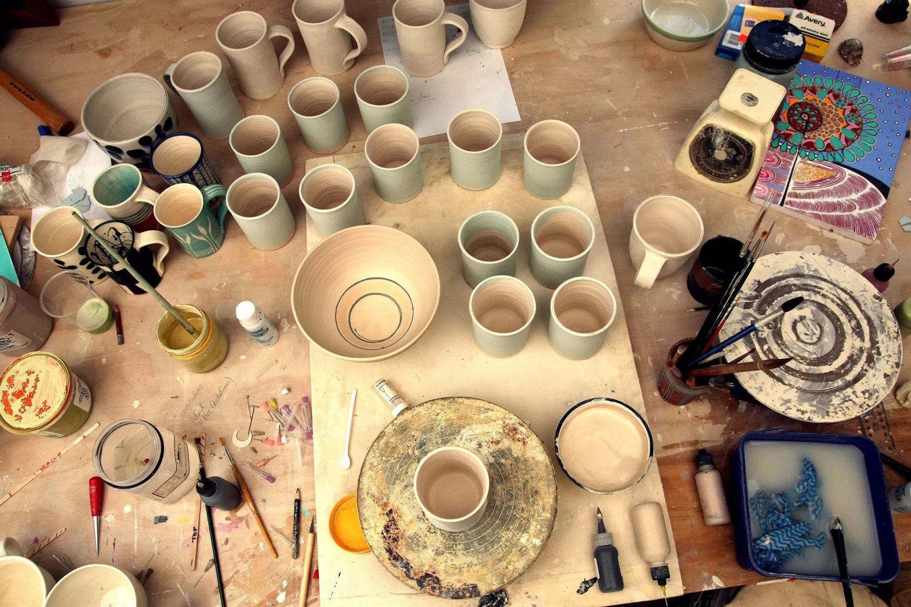 A cluttered pottery workspace with multiple ceramic cups, bowls, and pottery tools spread across a wooden table. Various paints, brushes, and scales are scattered around, along with colorful patterned artwork in the top right corner.