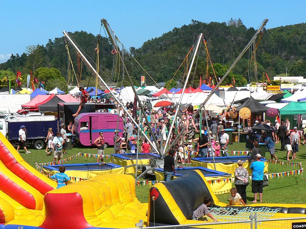 A vibrant fairground scene with a colorful bouncy castle in the foreground, a trampoline setup at the center, and numerous people enjoying the event. Tents and stalls are in the background, with trees and a clear blue sky completing the lively atmosphere.