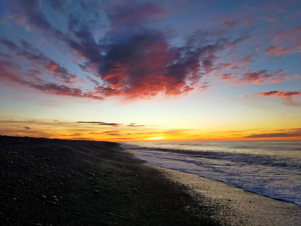 A serene beach scene at sunset, with the sky displaying a gradient of colors from deep blue to vibrant orange and pink. Gentle waves lap against the shore, and fluffy clouds catch the last light of day, creating a peaceful and picturesque moment reminiscent of a winter adventure by the sea.