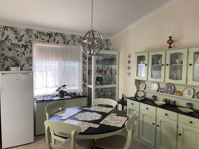 A cozy dining room with a round black table surrounded by light green chairs. A matching light green buffet hutch displays various dishware. The walls are adorned with floral-patterned wallpaper, and a modern pendant light hangs from the ceiling.