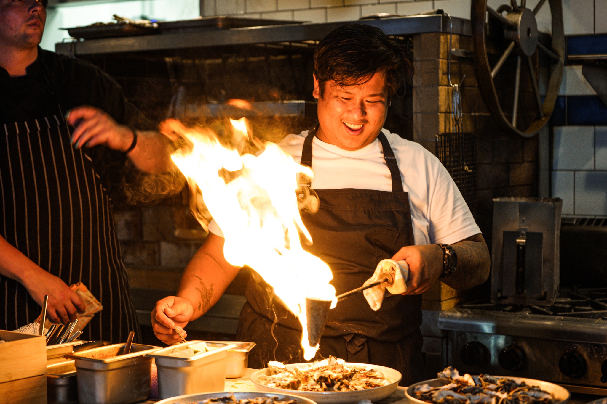 A chef in a white shirt and dark apron is energetically torching food on a plate, creating a large flame. He is smiling and surrounded by various kitchen equipment and dishes. Another person in a striped apron is partially visible in the background.